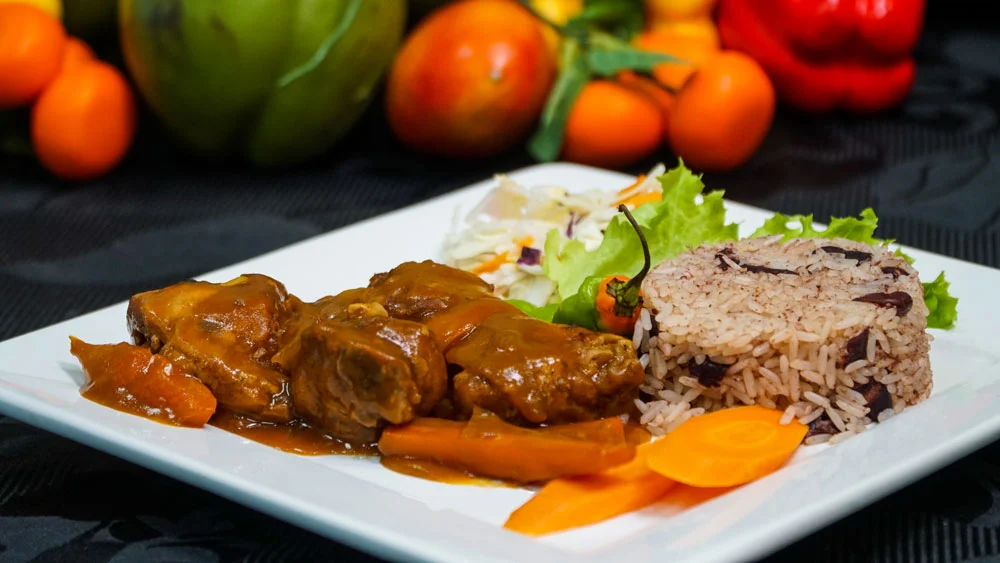 A captivating image showcasing the culinary excellence of Jerk and BBQ Haven, a premier West Indian restaurant near you. The photograph displays a delectable Oxtail Dinner, accompanied by flavorful Rice & Peas and a fresh Salad. 